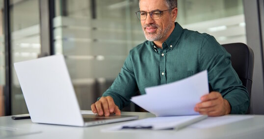 Smiling middle aged executive, mature male hr manager holding documents using laptop looking at pc computer in office at desk, checking financial data in report, doing account paper plan overview.